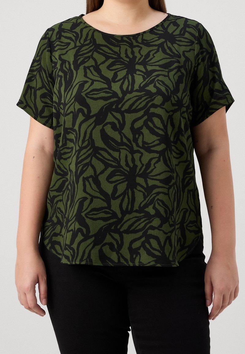 Femme portant un haut vert à manches courtes avec des motifs abstraits de feuilles noires et un pantalon noir, debout devant un fond uni.