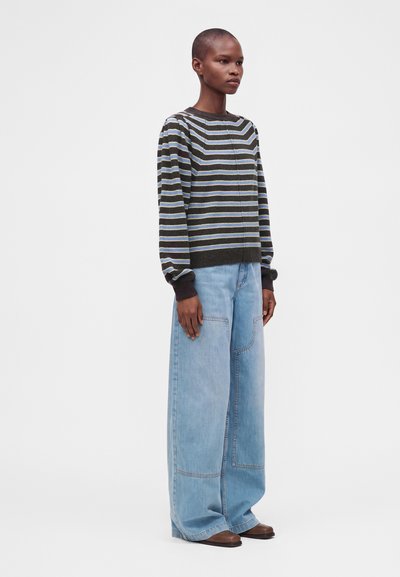Personne portant un pull rayé foncé et un jean bleu clair à jambes larges, debout devant un fond uni clair.