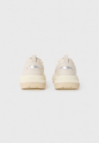 Beige sneakers with soft fabric, a thick cream sole, and metallic silver accents on the heel. Features a pull tab for easy wear.