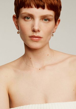 Se muestran un collar de estrellas doradas y unos pendientes circulares dorados; la modelo tiene el cabello corto y lleva una blusa blanca sin tirantes y sin patrones.