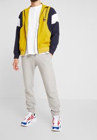 Chaqueta amarilla y azul marino con cremallera y logo, camiseta blanca con estampado, pantalones de chándal grises y zapatillas multicolor. Ropa deportiva casual.