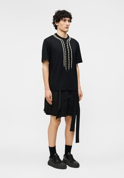 Νεαρός άνδρας που στέκεται, φορώντας μαύρο κοντομάνικο πουκάμισο με κορδόνια σχέδιο, μαύρη φούστα με στρώσεις, μαύρες κάλτσες και μαύρα αθλητικά παπούτσια.