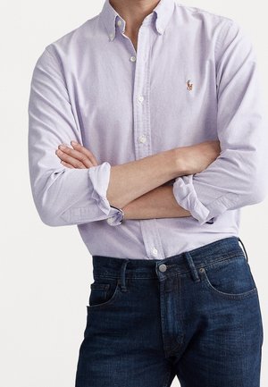Homme portant une chemise boutonnée violet clair à manches retroussées et un jean bleu foncé, debout les bras croisés.