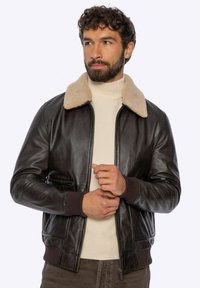 Bruine leren bomberjack met een beige shearlingkraag. Voorzien van een ritssluiting en geribbelde manchetten. Model draagt een crème coltrui eronder.