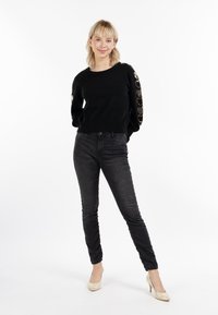 Pullover negru din tricot cu accente decorative aurii în formă de inimă pe mâneci, asociat cu jeansi negri skinny și pantofi cu toc înalt bej.