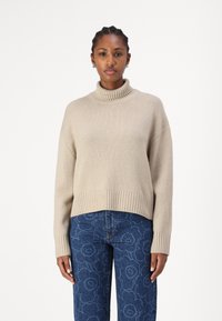 Beige Rollkragenpullover mit geripptem Saum und Bündchen, lockere Passform. Kombiniert mit blauen Jeans im Blumenmuster.