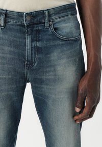 Jeans de mezclilla azul deslavado con corte recto, diseño de cinco bolsillos y detalles de remaches metálicos. La textura es suave con ligero desgastado.