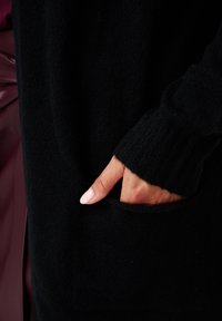 Hand met lichtroze nagels rustend in de zak van een zwarte gebreide trui.