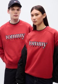 Κόκκινα φούτερ με το λογότυπο "tommy JEANS" σε αντίθετο λευκό χρώμα, με ριμπ μανσέτες και στρογγυλές λαιμόκοψεις. Μερικώς εμφανές μαύρο μπουφάν.