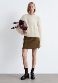 Cremefarbener, flauschiger Pullover, olivgrüne Rock mit Taschen, braune Slipper und gemusterte Socken, hält eine burgunderfarbene Tasche.