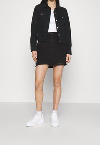 Giacca di jeans nera con dettagli a bottoni, abbinata a una gonna di jeans nera a vita alta con bordi sfilacciati. Sneakers bianche completano l'outfit.