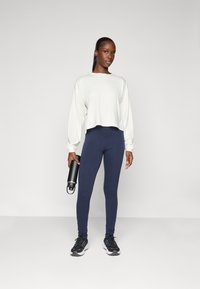 Balti trumpas ilgomis rankovėmis marškinėliai su nukritusiomis pečių siūlėmis, derinami su tamsiai mėlynomis leggingais. Modelis laiko juodą vandens butelį ir dėvi juodus sportbačius.