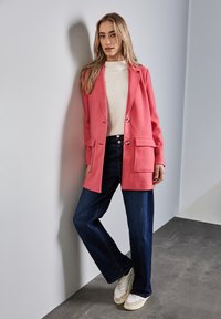 Blazer oversize in misto lana rosa con rever, due tasche frontali, abbinato a jeans a gamba larga blu scuro e sneakers bianche. Texture liscia.