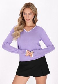 Lila V-Ausschnitt-Pullover mit langen Ärmeln, verziert mit weißen Akzenten am Halsausschnitt und an den Bündchen, kombiniert mit einem schwarzen, gefällteten Minirock.