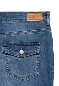 Blå denim jeans med et lærlabel, en klaff baklomme med en rund knapp og kontrasterende sømmer på lommekantene.