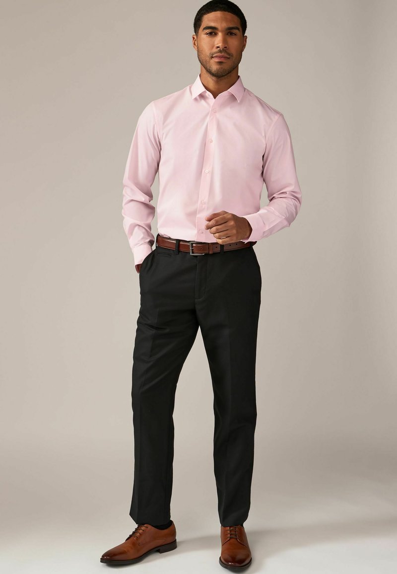 Camisa de vestir rosa claro con cuello, cierre de botones y mangas largas, combinada con pantalones negros y zapatos de cuero marrón.
