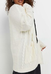 Chunky Strickjacke in Creme mit strukturierten Kabelmustern, lockerer Passform, rippenförmigen Bündchen und zwei großen Vordertaschen. Aus weichem und leichtem Material.