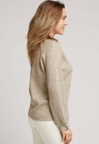 Pull en tricot beige à manches longues, avec des poignets côtelés et des accents de boutons. Texture douce, coupe décontractée, design sobre, porté avec un pantalon blanc.