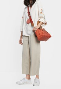 Kvinna klädd i krämfärgad blommig blus och beige byxor, hållande en texturerad orange handväska och en rund plånbok med matchande rem, iförd vita sneakers.