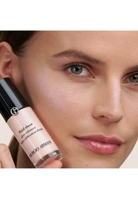 Fluid Sheer de Giorgio Armani en una botella de vidrio, presenta un líquido rosa claro, tapa negra y un diseño elegante. Sostenido junto a la piel con un brillo sutil.