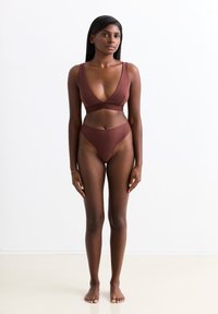 Vrouw die blootsvoets tegen een witte achtergrond staat, gekleed in een bruin diepe V-hals bralette en hooggeplaatste onderbroek, recht vooruit kijkend met ontspannen armen.