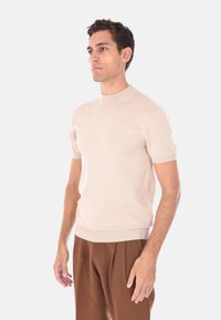 Uomo in piedi rivolto a sinistra, che indossa una maglietta a maniche corte beige in maglia e pantaloni marroni a pieghe su uno sfondo chiaro e uniforme.