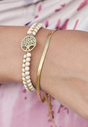 Weißes Perlenarmband mit einem goldenen Baum-des-Lebens-Akzent, kombiniert mit einem dünnen goldenen Bangle mit strukturiertem Oberflächenfinish.