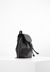 Czarny plecak ze skóry o zaokrąglonym kształcie, wyposażony w klapę, sznurek ściągający oraz srebrny łańcuszkowy pasek. Materiał ma teksturowane wykończenie.