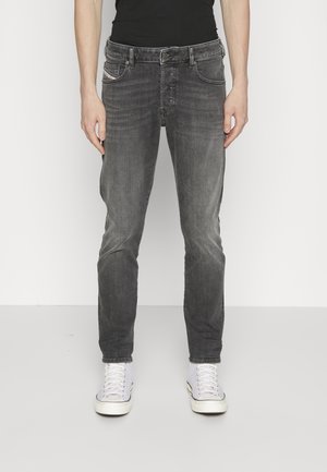 Personne portant un jean noir délavé slim-fit et des baskets montantes blanches, debout devant un fond blanc uni.