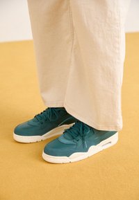 Baskets bleu sarcelle avec semelles blanches portées avec un pantalon large beige clair sur un sol jaune moutarde.