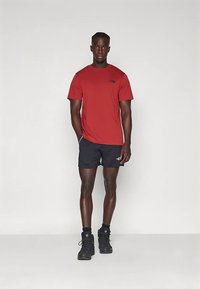 Homem em pé com as mãos nos bolsos, vestindo uma camisa de manga curta vermelha, calções pretos, meias pretas e botas de caminhadas pretas sobre um fundo liso.