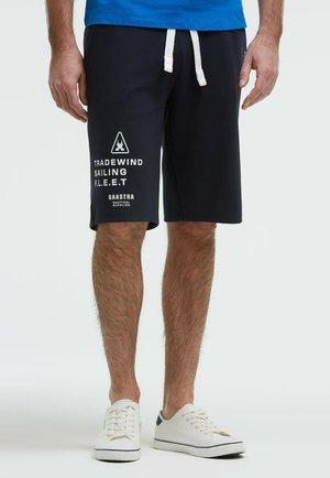 Mann trägt schwarze Segelshorts mit weißen Kordeln und nautischem Schriftzug, weiße Turnschuhe und ein teilweise sichtbares blaues Hemd.