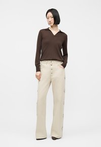 Γυναίκα που στέκεται με το ένα χέρι στην τσέπη, φοράει καφέ μπλούζα με μακριά μανίκια και μπεζ παντελόνι με φαρδιά μπατζάκια και πλευρικές λεπτομέρειες κουμπιών.