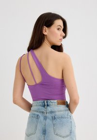 Top de tirantes ajustado en color púrpura con diseño asimétrico y múltiples tiras finas, combinado con pantalones cortos de mezclilla claros de tiro alto que cuentan con bolsillos traseros.