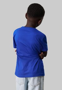 T-shirt in cotone blu con maniche corte, colletto tondo e una texture liscia. La vista posteriore mostra un design semplice, senza grafiche o accenti.