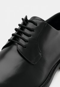 Chaussure habillée en cuir noir avec une finition lisse, dotée de lacets droits et d'un embout rond. Design minimaliste sans ornements visibles.