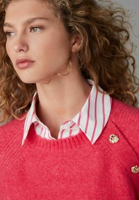 Lyserød sweater med ribber i halsudskæringen, prydet med gyldne blomsterknapper. Båret over en hvid skjorte med lyserøde striber, der fremhæver lagdelte teksturer.