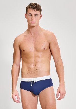 Bărbat tânăr cu păr scurt purtând slip de baie bleumarin cu talie albă și șnur, stând în picioare pe un fundal simplu și deschis la culoare.