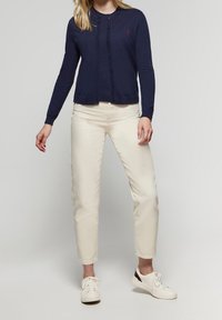 Námořnický kardigan na knoflíky s kulatým výstřihem, v kombinaci s krémovými zužujícími se kalhotami a bílými teniskami. Kardigan má na hrudi malý emblém.