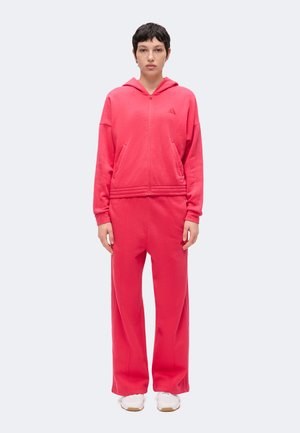 Personne debout face à l'objectif portant un sweat à capuche rouge zippé et un pantalon large assorti avec des baskets blanches sur un fond uni.