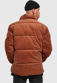 Brun kordfløyels pufferjakke med horisontal quiltet design, høy krage og teksturert stoff. Ingen synlige maskinvare eller festemidler.