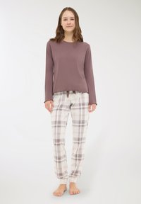 Giovane donna in piedi a piedi nudi, che indossa una maglietta a maniche lunghe viola tenue e pantaloni del pigiama a scacchi bianchi, su uno sfondo chiaro e semplice.