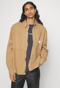 Giacca camicia in cotone color tan con bottoni a pressione, due tasche frontali e un piccolo patch con logo. Indossata sopra una maglietta scura, abbinata a pantaloni neri.