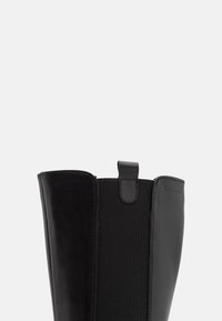 Gros plan sur la partie supérieure d'une botte en cuir noire avec un panneau latéral élastique et une languette de tirage au centre supérieur.