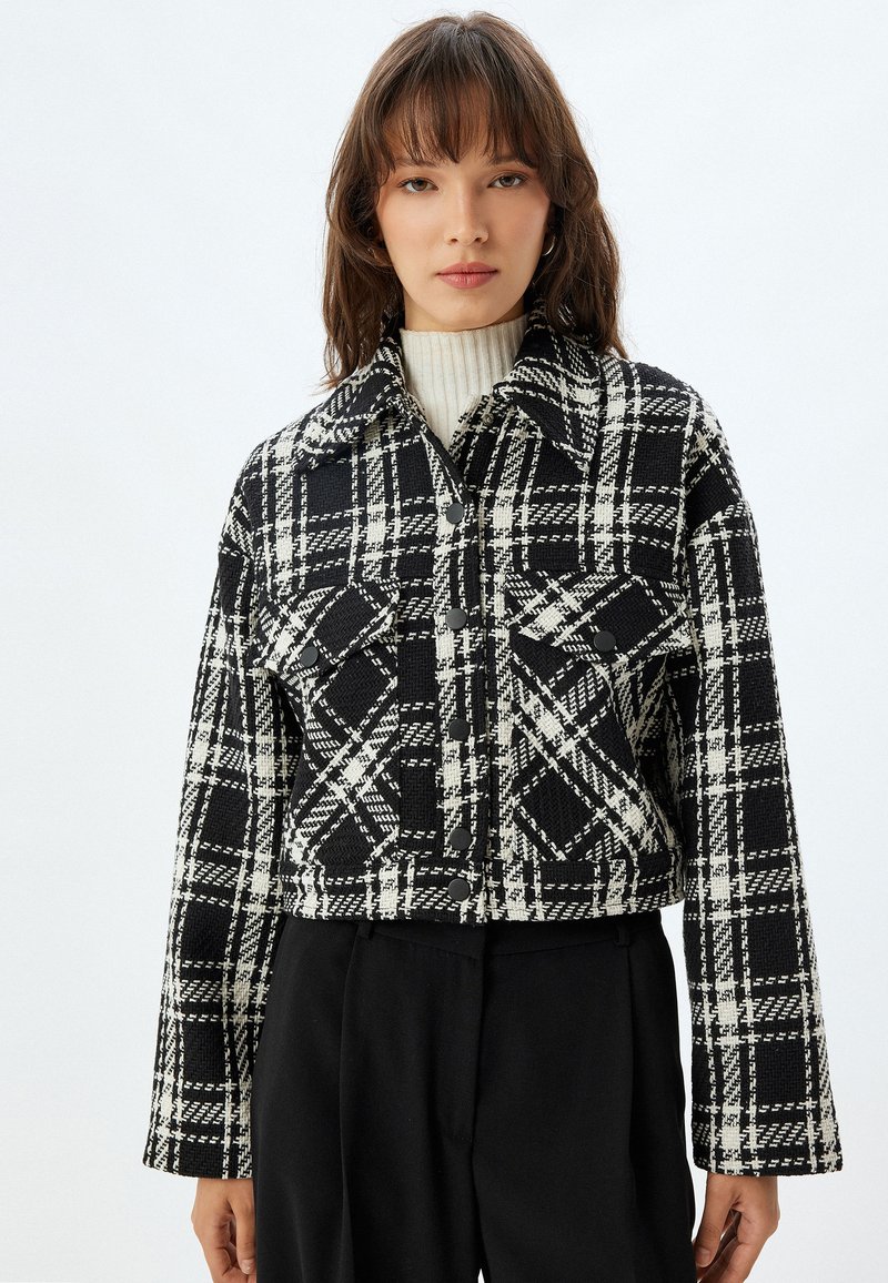 Chaqueta corta a cuadros en blanco y negro con gran collar, botones frontales y dos bolsillos en el pecho. Textura tejida con patrón de cuadros.