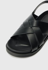 Black leather sandal with crossover straps, open heel, and a textured footbed. Features a flat, rounded sole and minimalist design.