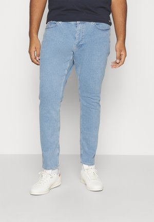 Homme portant un jean slim bleu clair, des baskets blanches et une chemise sombre, debout devant un fond gris clair uni.
