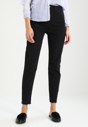 Jeans skinny negros con dobladillos deshilachados, hechos de denim. Llevados con una camisa azul clara de botones y unos zapatos de casa negros de felpa.