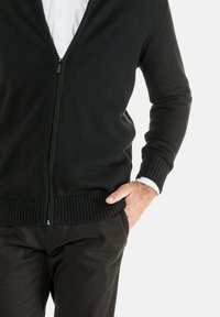 Schwarze, mit Reißverschluss versehene Strickjacke mit gerippten Bündchen und Saum, getragen über einem weißen Hemd. Der Stoff hat eine glatte Textur und zeigt ein schmal geschnittenes Design.