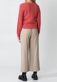 Röd kabelstickad tröja med ribbad nederkant, i kombination med beiga vidbyxor och svarta ankelskor. Slät textur och avslappnad passform.